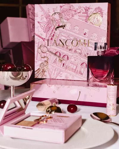 Unnepi Lancome holiday kollekcios display egy asztalon, nagy illusztralt rozsaszin ajandekdobozzal, parfummel, sminktermekekkel es friss cseresznyekkel.