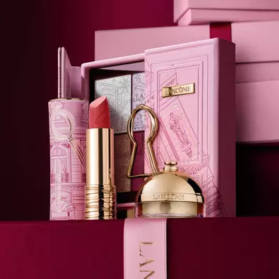 Gondosan osszeallitott Lancome unnepi ajandekszett ruzzsal, szemhejpalettaval es arany beauty termekekkel, dekorativ rozsaszin tok előtt elrendezve.