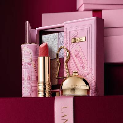 Gondosan osszeallitott Lancome unnepi ajandekszett ruzzsal, szemhejpalettaval es arany beauty termekekkel, dekorativ rozsaszin tok előtt elrendezve.