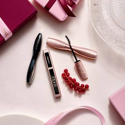 Lancome sminktermekek flat lay elrendezese szempillaspirallal es szajfennyel, rozsaszin ajandekdobozokkal, szatenszalagokkal es egy voros ribizliaggal.