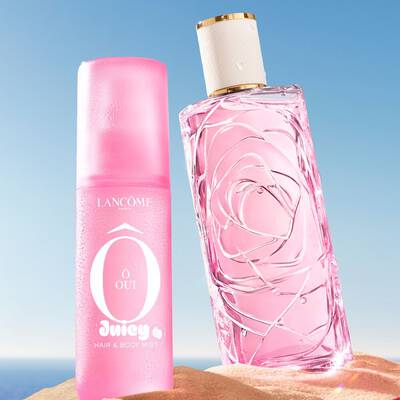 Egy tengerparti jelenet, ahol harom szines parfumuveg all egyenesen a homokban, egy rozsaszin, egy sarga, egy zold, mig egy masik rozsaszin parfum uveg az oldalan fekszik az előterben. Egy rozsaszin-feher csikos torolkozőt helyeznek el a homokra bal oldalon. A hatterben tiszta kek egbolt alakul at a horizonton.
