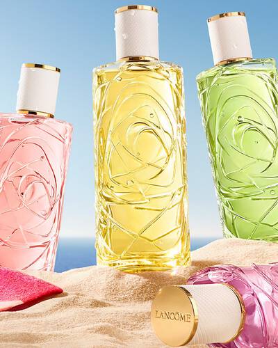 Ket rozsaszin uveg, egy parfum, atlatszo uveg texturalt mintaval, es egy permetező uveg, homokos tengerparton allva. Egy osszehajtott, rozsaszin csikos strandtorlő fekszik az uvegek mellett. A hatterben tiszta kek eg es az ocean lathato.