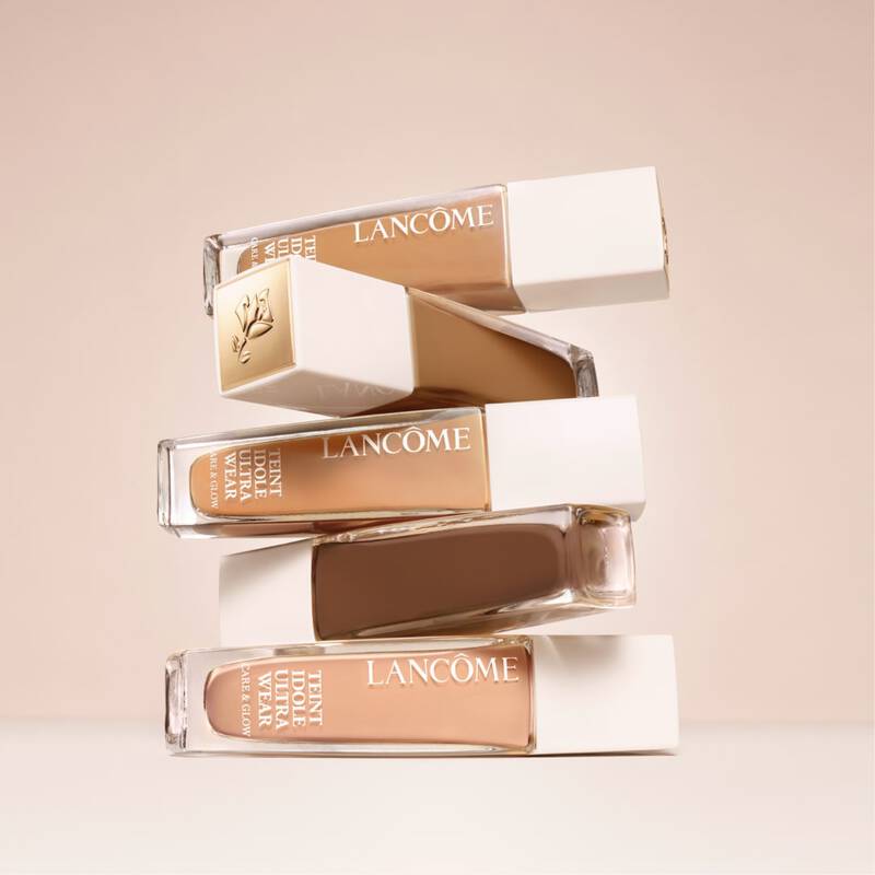 A stack of four Lancome foundation bottles in various shades, featuring a clean, minimalist design against a soft, neutral background.