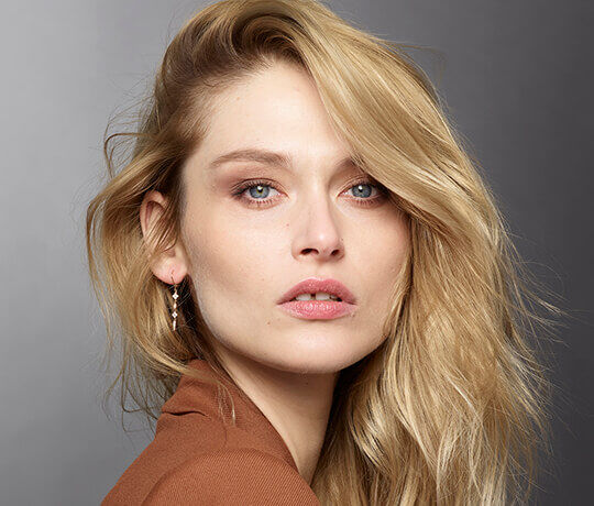 A woman with long, wavy blonde hair and blue eyes gazes directly at the camera, wearing subtle makeup and dangling earrings. She has a neutral expression and is dressed in a brown top against a gray background.