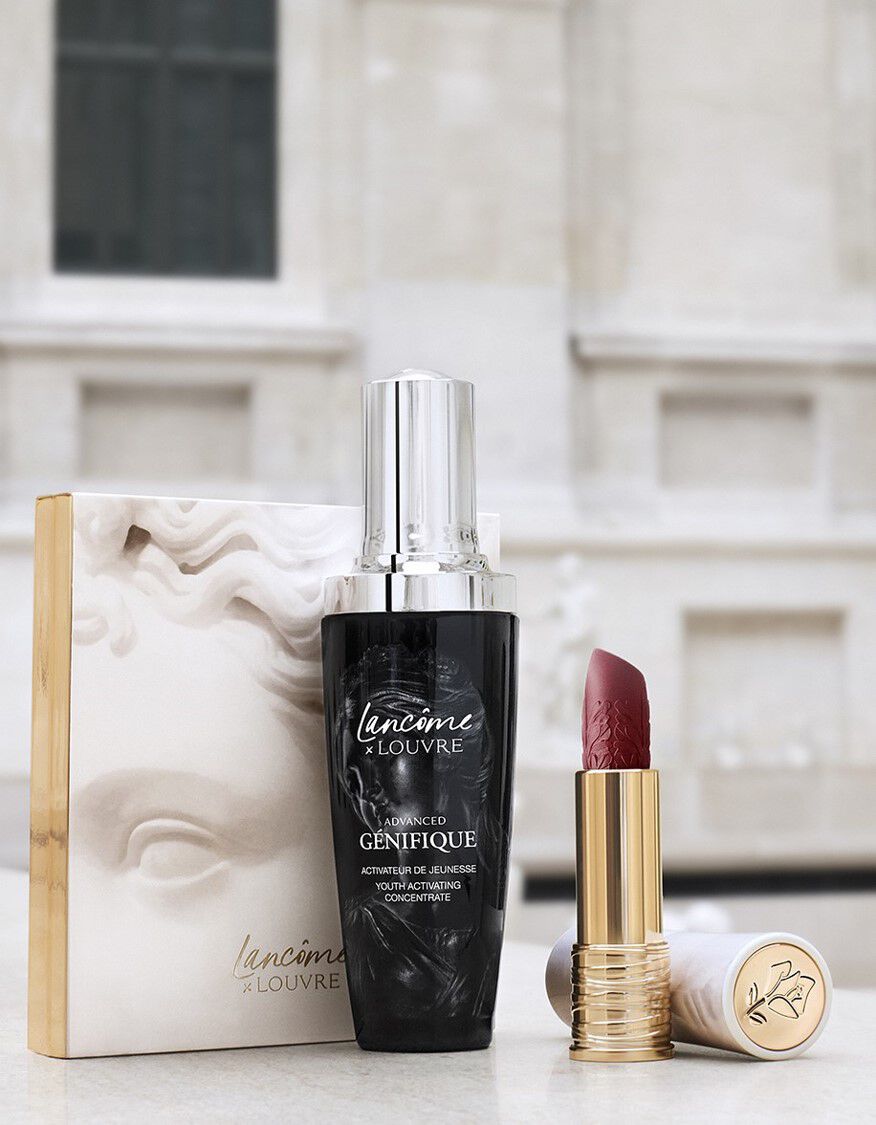 A bottle of Lancome Advanced Genifique serum and a red lipstick in a gold casing, displayed alongside an elegant box featuring a sculpted design, set against a blurred background of a classic architectural structure.