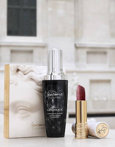 A bottle of Lancome Advanced Genifique serum and a red lipstick in a gold casing, displayed alongside an elegant box featuring a sculpted design, set against a blurred background of a classic architectural structure.