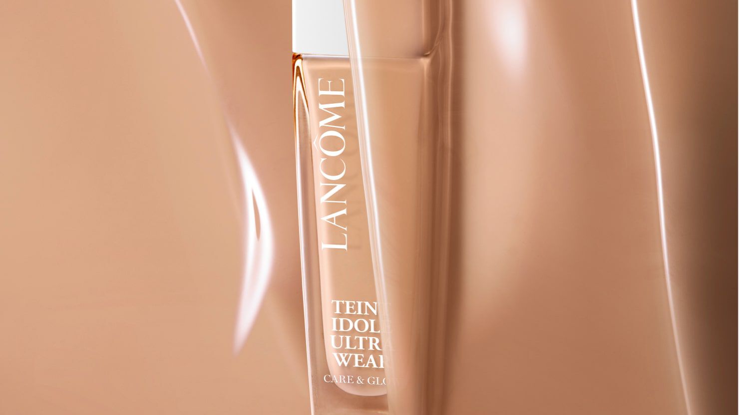 A close-up of a Lancome foundation bottle partially obscured in a warm-toned background. The bottle is sleek and elegant, labeled "Teint Idole Ultra Wear."