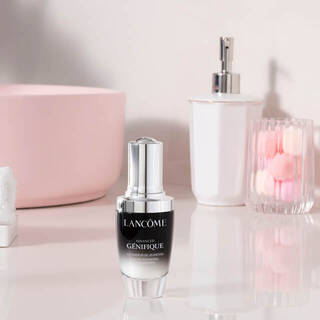 A Lancome Genifique serum bottle displayed on a reflective surface, accompanied by pink flowers and elegant glass vases in a soft pastel setting.