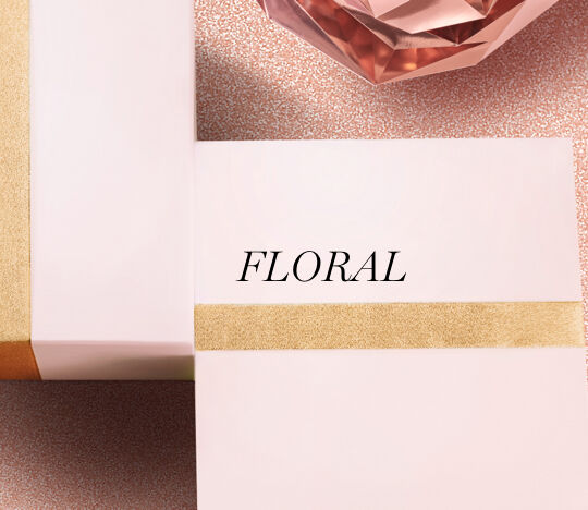 A close-up of two elegantly wrapped gift boxes with a gold stripe, featuring the word "FLORAL" in bold black letters. In the background, there's a decorative gem in a soft pink hue, set against a textured, sparkly surface.