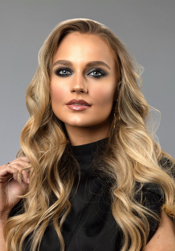 A model with long, wavy blonde hair and smoky eye makeup poses confidently against a neutral background, wearing a black top.