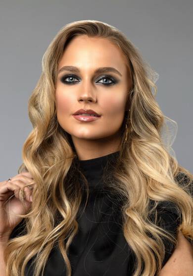 A model with long, wavy blonde hair and smoky eye makeup poses confidently against a neutral background, wearing a black top.