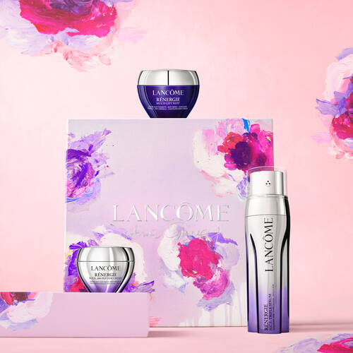 Alt text: A product display featuring Lancome skincare items, including two jars and a bottle, on a pastel pink background with colorful floral accents. The packaging is beautifully designed with the Lancome logo.