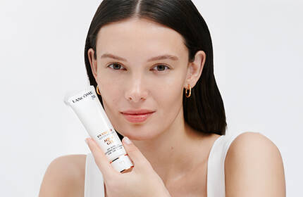 A woman with long, dark hair holds a skincare device while smiling at the camera against a light background.