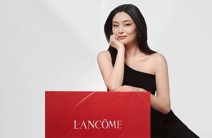 A woman in a black dress rests her chin on her hand while sitting beside a large red Lancome box against a light background.