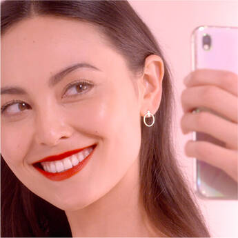 A woman with long dark hair smiles brightly while taking a selfie, wearing bold red lipstick and hoop earrings. She is in a softly lit environment with pastel colors in the background.