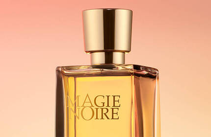 A glass perfume bottle with a gold cap, displaying the label "MAGIE NOIRE," against a soft gradient background in warm shades.