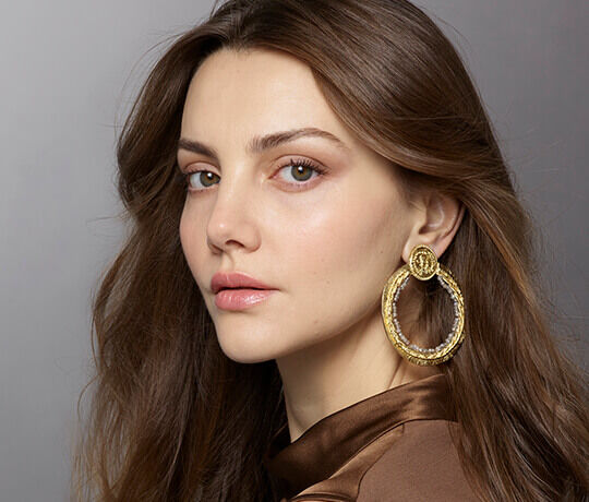 A close-up portrait of a woman with long, wavy brown hair, wearing large circular gold earrings. She has a natural makeup look and is gazing slightly to the side.