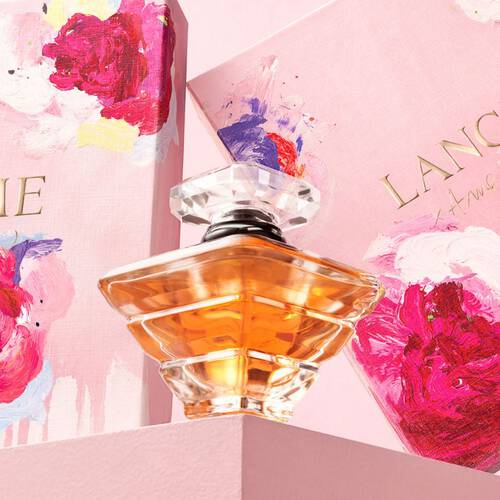 A close-up of a glass perfume bottle with a crystal cap, placed alongside beautifully designed pink boxes featuring floral artwork. The bottle contains a golden liquid, and the overall composition has a delicate and elegant feel.
