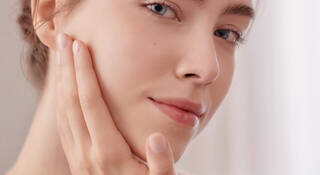 A young woman gently touching her face, with a soft expression and natural makeup, looking directly at the camera.