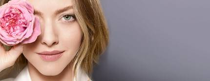 A woman with light brown hair holds a pink rose to her eye, smiling softly against a muted gray background.