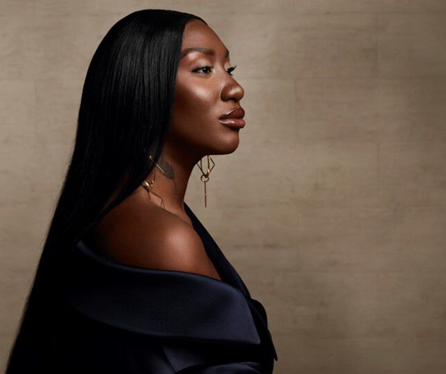 A profile portrait of a woman with long, straight black hair, wearing an off-the-shoulder dark garment. She has bold makeup and is adorned with elegant earrings, set against a neutral background.