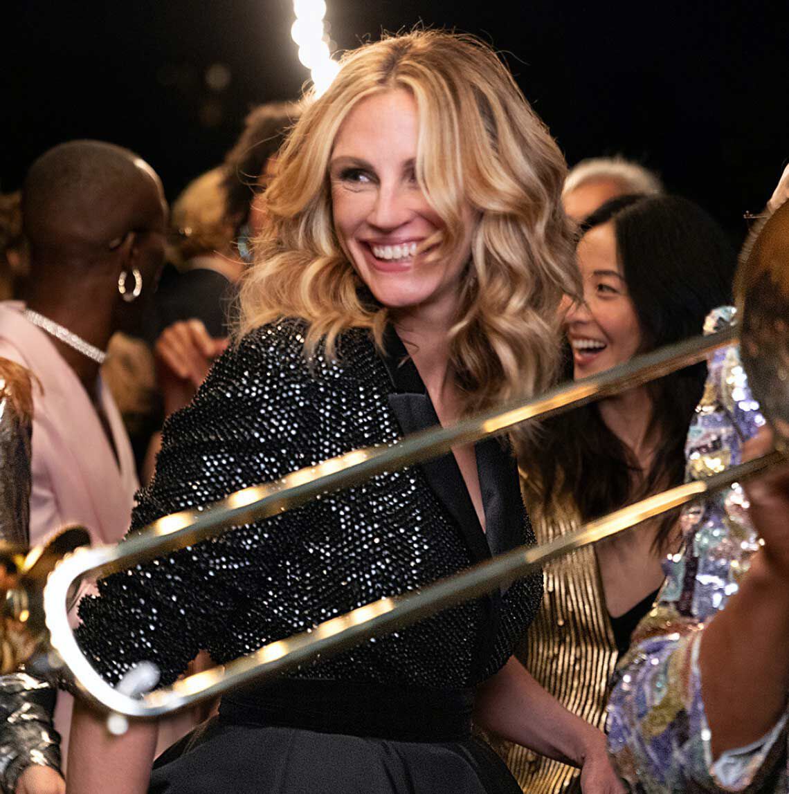 Egy elegans nő, hosszu, hullamos hajjal, vidaman mosolyog egy unnepi esemenyen, mikozben trombita jatekosok es mas vendegek veszik korul.