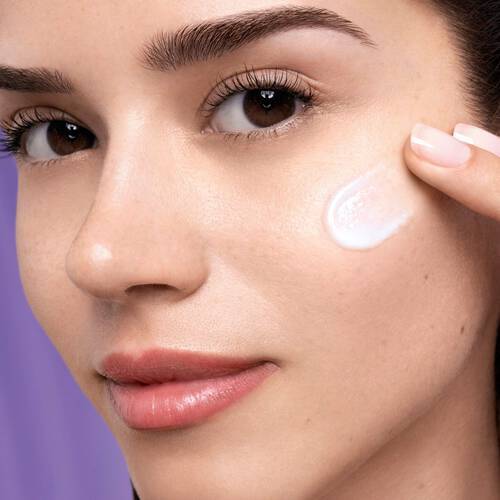 Egy fiatal nő a bőrere kremet visz fel, kozelről nez a kameraba, termeszetes sminkkel es szep bőrrel.