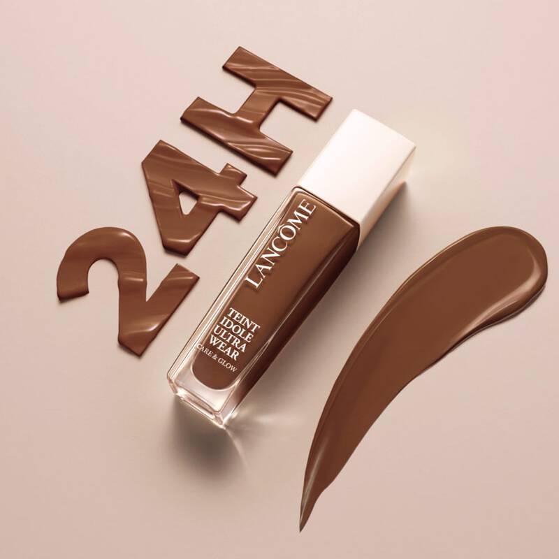 A bottle of Lancome foundation in a deep brown shade, with a swatch of the product beside it and the text "24H" made from chocolate-colored material in the background.