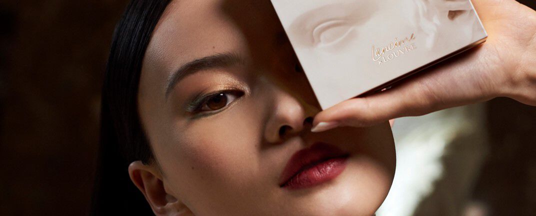 A close-up of a model's face, showcasing elegant makeup with bold lips, as she holds a skincare product box near her face.