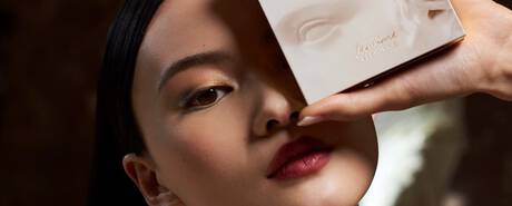 A close-up of a model's face, showcasing elegant makeup with bold lips, as she holds a skincare product box near her face.