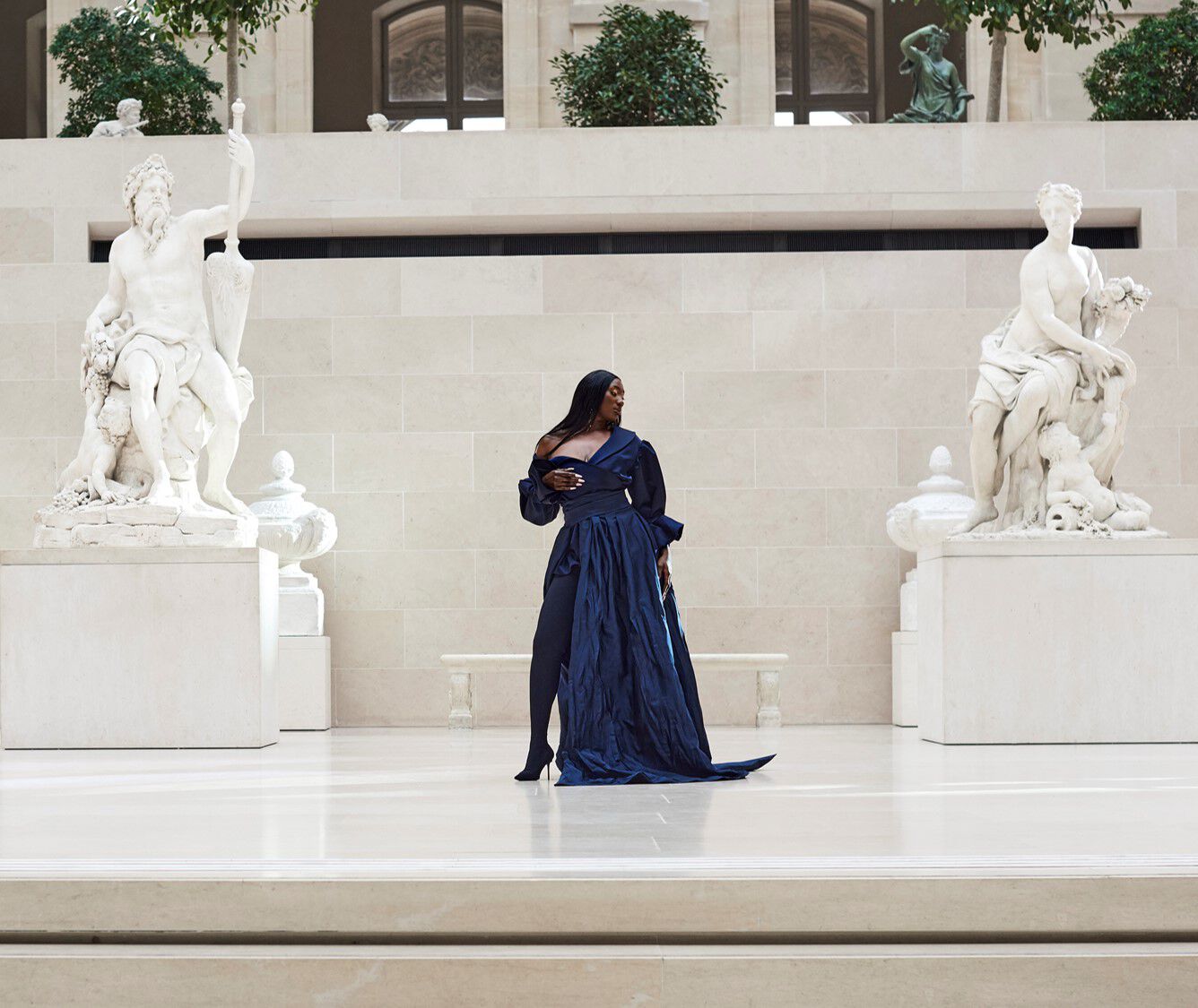 A model stands gracefully in a spacious gallery, wearing a flowing navy blue gown, with classical sculptures in the background.